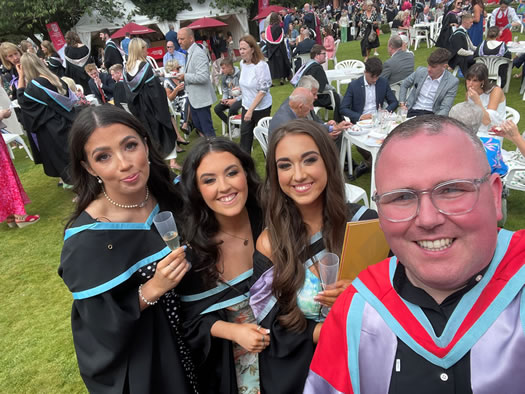 a selfie photo taken by Dr Thomas Rogan, a lecturer at St Mary's with three of the Irish Medium graduates of the day.  The three girls behind him are smiling, wearing their graduation gowns and the further background shows outside on the grass, lots of the graduates, their family and friends all enjoying some refreshments after the ceremony.